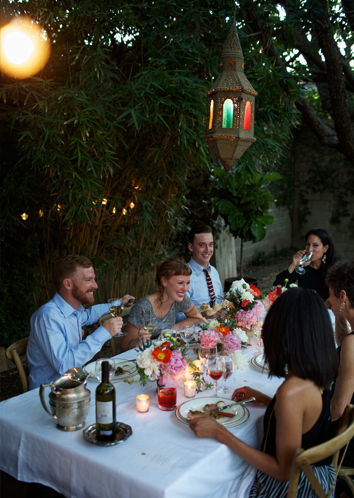 A picture of friends dining at a white table decorated with white, orange and pink flower bouquets.