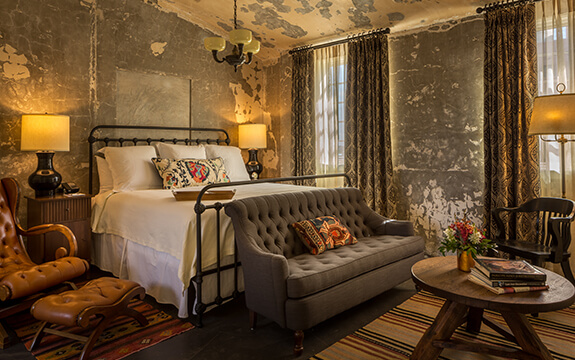 A picture of Hotel Emma's version of a presidential suite, named the Emma Koehler suite. Pictured is the tall ceiling room with king-sized white bed with a grey couch in front of it and a tan chair next to it.