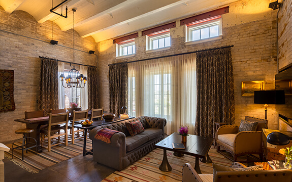 A picture of Hotel Emma's Maritzen suite. Pictured is the living room with a spacious seating arrangement, a unique 8 person table and windows with curtains.